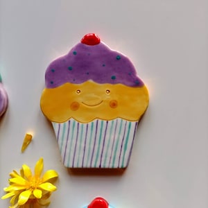 May include: A ceramic cupcake with purple frosting, yellow cake, and a red cherry on top. The cupcake has a happy face and is decorated with green and purple stripes.