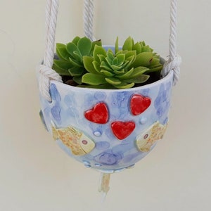 May include: A blue ceramic hanging planter with a white rope, decorated with red hearts and yellow fish. The planter is filled with green succulents.