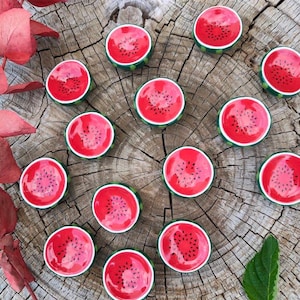 Può includere: Una collezione di oggetti decorativi a tema anguria. Ogni oggetto è rotondo con interno rosso, dettagli di semi neri e scorza verde. Disposti su una superficie di legno stagionato, con foglie rosse e una foglia verde.
