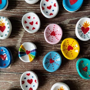 Puede incluir: Una colección de pequeños cuencos de cerámica pintados a mano en varios colores, incluyendo blanco, azul, amarillo y rosa. Cada cuenco presenta un diseño de corazón, algunos con llamas o patrones de puntos. Los cuencos están dispuestos sobre una superficie de madera.