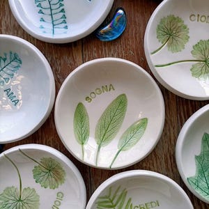 Può includere: Una collezione di piccole ciotole in ceramica bianca con disegni botanici e parole italiane. Le ciotole presentano motivi di foglie e felci verdi, con parole come "AMORE" e "SOGNA". Un elemento decorativo in vetro blu è presente tra le ciotole.