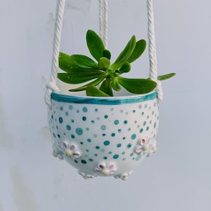 Pode incluir: Um vaso de cerâmica branco pendurado com um padrão de bolinhas verdes e flores brancas ao redor da borda. Uma pequena planta suculenta verde está crescendo dentro do vaso.