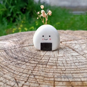 May include: A white ceramic onigiri-shaped vase with a smiling face and pink cheeks. The vase holds a small bouquet of dried flowers. The vase is placed on a weathered wooden surface, with a blurred green background.