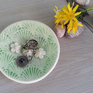 Könnte beinhalten: Eine grüne Keramikschale mit einem fächerförmigen Design und weißen Blumen. Die Schale ist gefüllt mit Schmuck, darunter ein silberner Ring, ein silbernes Armband mit Charms und ein silberner Anhänger mit einem blauen Auge.