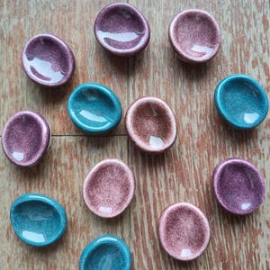 Può includere: Una collezione di piccoli piatti in ceramica di forma ovale nei toni del viola, rosa e verde acqua. I piatti hanno una finitura lucida e macchiettata e sono disposti su una superficie di legno. L'estetica generale è minimalista e artistica.