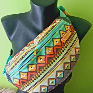 Peut inclure: Sac banane coloré avec un motif géométrique dans les tons de turquoise, orange, jaune et marron. Le sac a une fermeture éclair et une sangle turquoise, et une petite étiquette en cuir qui dit "HANDMADE LIMITED EDITION".