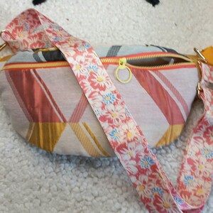 Peut inclure: Une banane à motif floral rose et blanc avec une fermeture éclair dorée et une bandoulière assortie. Le sac a un motif géométrique dans des tons de rose, or et blanc.