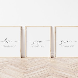 May include: Three framed prints with white backgrounds and black calligraphy. The prints read "love is spoken here", "joy is chosen here", and "grace is given here".