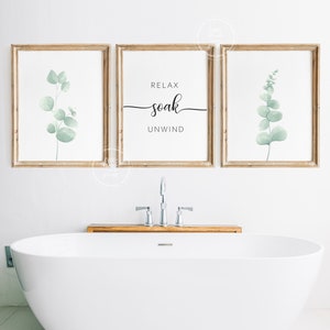 May include: Three framed prints with a white background and wooden frames. The center print has the text "Relax soak unwind" in black cursive font. The left and right prints feature a watercolor illustration of a green eucalyptus branch.