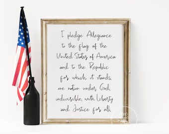 May include: A framed print of the Pledge of Allegiance in black cursive text on a white background. The frame is a light wood color. A small American flag and a black bottle are on a white surface to the left of the frame.