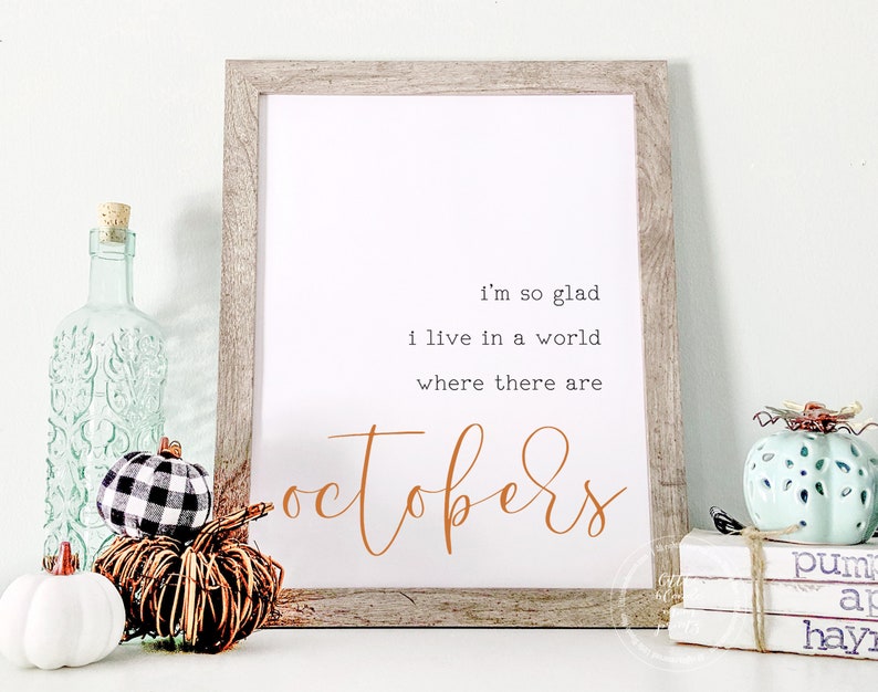 May include: A framed print with the text "i'm so glad i live in a world where there are octobers". The print is on a white background with brown text. The frame is a light wood color. There are small decorative pumpkins in the foreground.