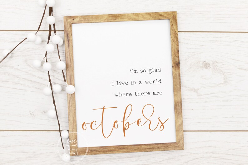 May include: A framed print with a white background and brown wood frame. The print features a quote in black text that reads "I'm so glad I live in a world where there are Octobers." The word "Octobers" is written in a cursive font in orange.