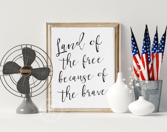 May include: A framed print with the text "Land of the free because of the brave" in black cursive script on a white background. The print is framed in a light wood frame and is sitting on a white surface with a fan, vases, and small American flags.