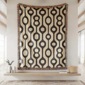 May include: A rectangular woven throw blanket featuring a repeating geometric pattern of dark charcoal circles and elongated ovals on a cream background. The blanket has a decorative fringe along the top and bottom edges with multicolored threads.