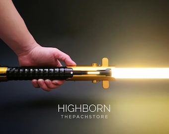 May include: A gold lightsaber with a black handle and a bright yellow blade. The lightsaber is lit and the blade is extended. The text "HIGHBORN THEPACHSTORE" is visible below the lightsaber.