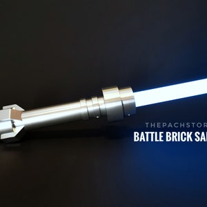 May include: A silver lightsaber with a blue blade. The lightsaber is made of metal and has a textured surface. The text "THEPACHSTORE BATTLE BRICK SABER" is visible in the image.