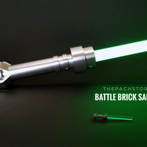 May include: A silver lightsaber hilt with a green blade. The hilt is made of metal and has a textured surface. The blade is about 2 feet long and is glowing brightly. The text "THEPACHSTORE BATTLE BRICK SABER" is visible below the lightsaber.