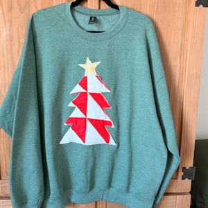 May include: A heather green crewneck sweatshirt featuring a central applique of a Christmas tree. The tree is constructed from alternating red and white fabric triangles and is topped with a single gold star. The garment has long sleeves.