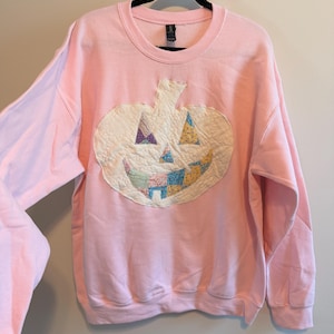 May include: A light pink crewneck sweatshirt featuring a large, central pumpkin appliqué. The pumpkin is constructed from quilted, off-white fabric with colorful, multi-patterned fabric patches forming the eyes, nose, and a wide, toothy grin.