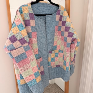 May include: A quilted jacket featuring a patchwork design of various floral and dotted fabrics in shades of pink, yellow, purple, and blue. The garment has a blue textured fabric center panel, a round neckline, and two front patch pockets.