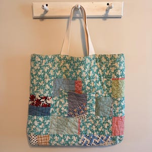 May include: A quilted tote bag constructed from a patchwork of various fabric scraps. The primary fabric features a teal floral pattern, interspersed with squares of red gingham, blue denim, striped, and polka-dot textiles. It has two cream-colored straps.