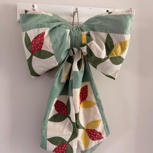 May include: A large decorative bow constructed from quilted fabric featuring a patchwork pattern of sage green borders and floral motifs in red, yellow, and dark green on an off-white background, hanging from a white wooden wall hook.