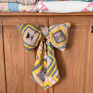 May include: A large decorative bow crafted from vintage patchwork quilt fabric featuring a geometric pattern in shades of yellow, grey, pink, and white. The bow has two wide loops and two long, trailing tails, all constructed from stitched fabric squares.