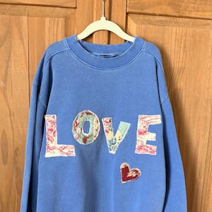 May include: A light blue crewneck sweatshirt featuring the word LOVE appliqued in various floral patterned fabric patches across the chest, accompanied by a small, dark red heart-shaped fabric patch positioned below the letters.