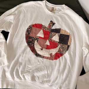 May include: A white long-sleeve crewneck sweatshirt featuring a large, central pumpkin applique made from a patchwork of various red, brown, and floral fabric squares. The pumpkin has white fabric eyes and a wide, grinning mouth.
