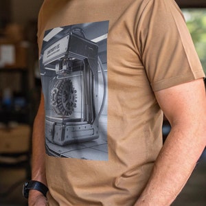 May include: A man wearing a brown t-shirt with a black and white image of a 3D printer on the front. The image shows the printer in action, with a gear in the centre of the print bed. The man is standing in a workshop setting.