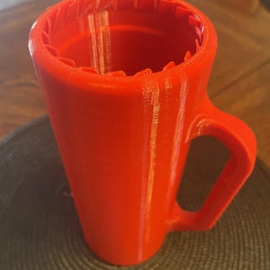 Puede incluir: Una taza de color rojo brillante con asa y borde texturizado. La taza tiene forma cilíndrica con una parte superior ligeramente ensanchada. El asa es un diseño de bucle simple. La taza está sobre un salvamanteles circular gris oscuro.