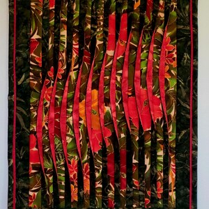 May include: A handmade textile art piece featuring a vibrant floral design. Vertical red fabric strips are layered over a background of green and brown floral fabric. The piece is framed with a dark green border and a thin red border.