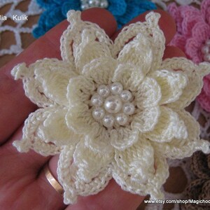 May include: A white crocheted flower with a pearl center. The flower is held in a hand with a ring on the finger.