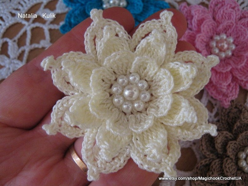 May include: A white crocheted flower with a pearl center. The flower is held in a hand with a ring on the finger.