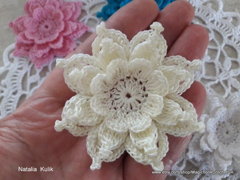 May include: A close-up of a crocheted white flower with a delicate, layered design. The flower is held in a hand, and there are other crocheted flowers in the background.