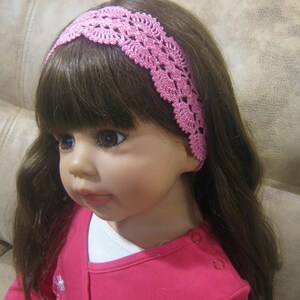 May include: A pink crocheted headband with a delicate floral pattern. The headband is worn by a doll with long brown hair.