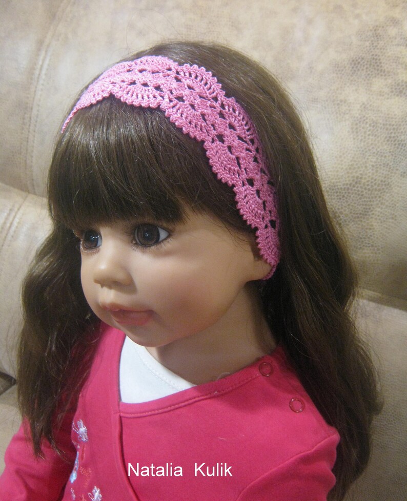 May include: A pink crocheted headband with a delicate floral pattern. The headband is worn by a doll with long brown hair.