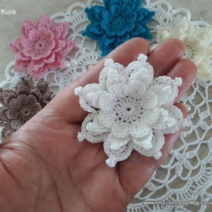 May include: A collection of crocheted flowers in various colors, including white, pink, brown, and blue. The flowers are arranged on a white crocheted doily.