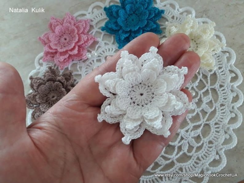 May include: A collection of crocheted flowers in various colors, including white, pink, brown, and blue. The flowers are arranged on a white crocheted doily.