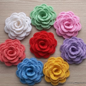 Könnte beinhalten: Eine Sammlung von acht gehäkelten Blumenapplikationen in verschiedenen Farben, darunter Weiß, Grün, Rosa, Rot, Lila, Blau und Gelb. Jede Blume hat eine spiralförmige Mitte und geschichtete Blütenblätter, ideal für Bastelprojekte.