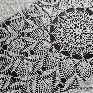 May include: A white, crocheted table runner with a detailed, openwork design. The runner features a central circular motif surrounded by repeating patterns of leaves and geometric shapes. The delicate lace-like texture is visible throughout the piece.