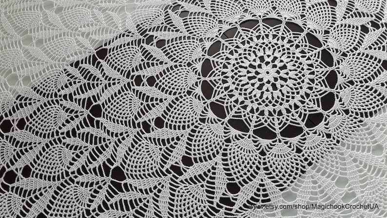 May include: A white, crocheted table runner with a detailed, openwork design. The runner features a central circular motif surrounded by repeating patterns of leaves and geometric shapes. The delicate lace-like texture is visible throughout the piece.
