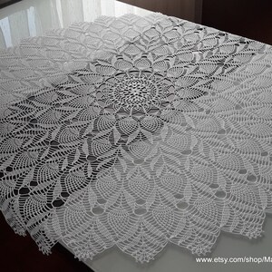 May include: A white, circular, crocheted tablecloth with a detailed floral pattern. The intricate design features a central motif that radiates outwards, creating a textured, decorative piece. The tablecloth is displayed on a table.