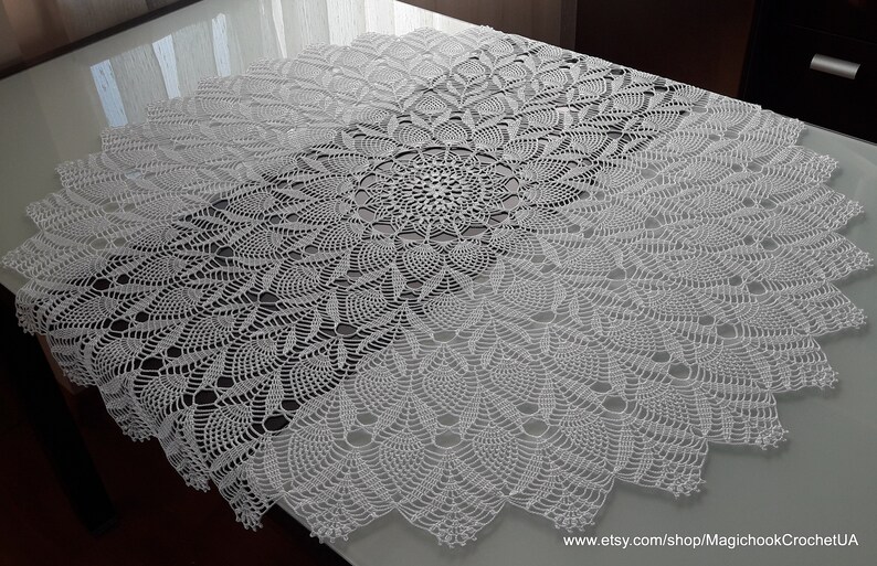 May include: A white, circular, crocheted tablecloth with a detailed floral pattern. The intricate design features a central motif that radiates outwards, creating a textured, decorative piece. The tablecloth is displayed on a table.