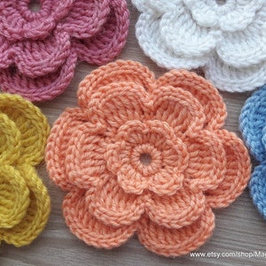May include: Five crocheted flowers in different colors, including pink, yellow, peach, white, and blue. The flowers are all made with a similar stitch pattern and have a layered, ruffled appearance.