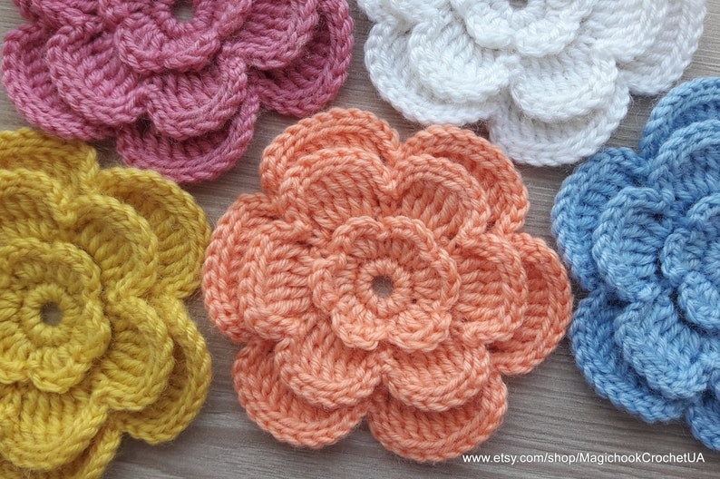 May include: Five crocheted flowers in different colors, including pink, yellow, peach, white, and blue. The flowers are all made with a similar stitch pattern and have a layered, ruffled appearance.