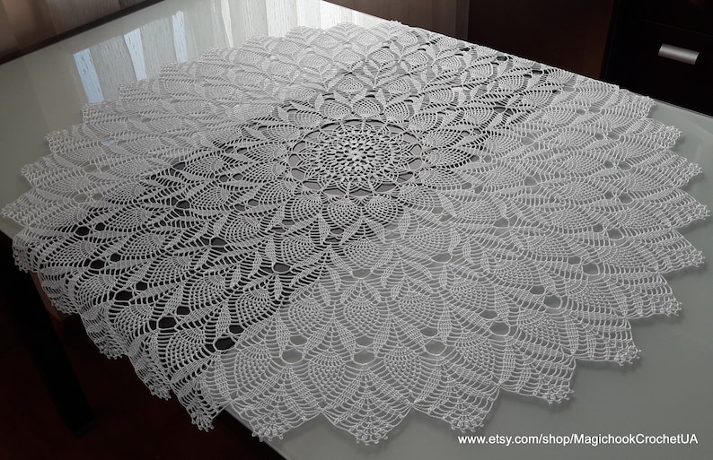 May include: A white, circular, crocheted tablecloth with a detailed floral pattern. The intricate design features a central motif that radiates outwards, creating a textured, decorative piece. The tablecloth is displayed on a table.