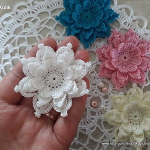 May include: Four crocheted flowers in white, pink, yellow, and blue. The flowers are arranged on a white crocheted doily. The flowers are all the same size and have a similar design.