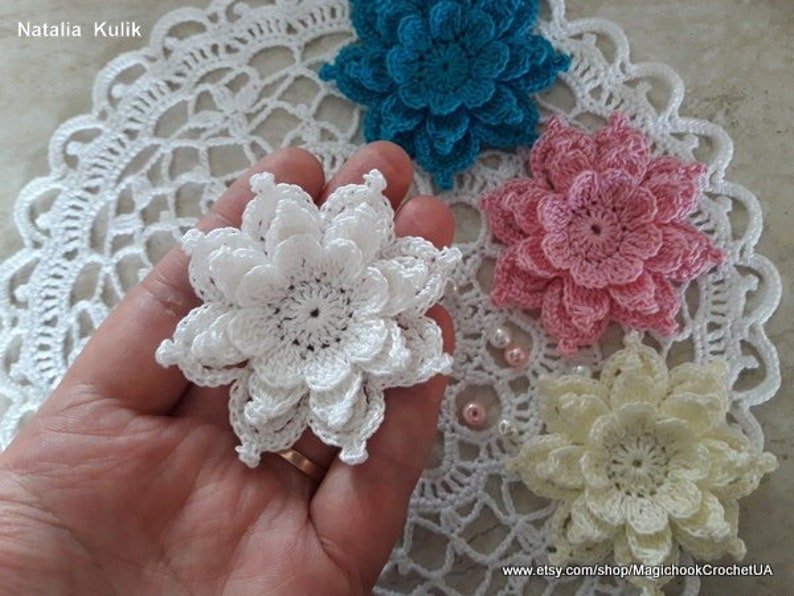 May include: Four crocheted flowers in white, pink, yellow, and blue. The flowers are arranged on a white crocheted doily. The flowers are all the same size and have a similar design.