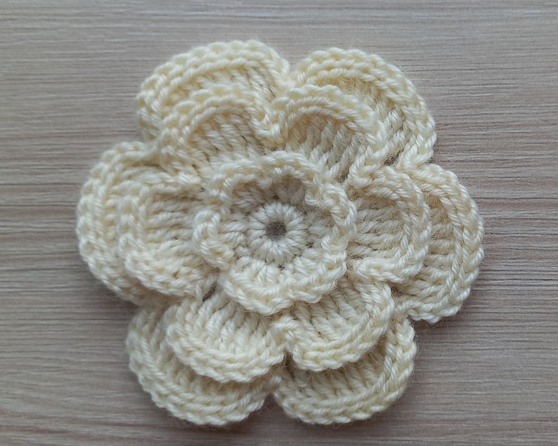 May include: A crocheted flower with six petals in a cream color. The flower is made with a single crochet stitch and has a center circle.